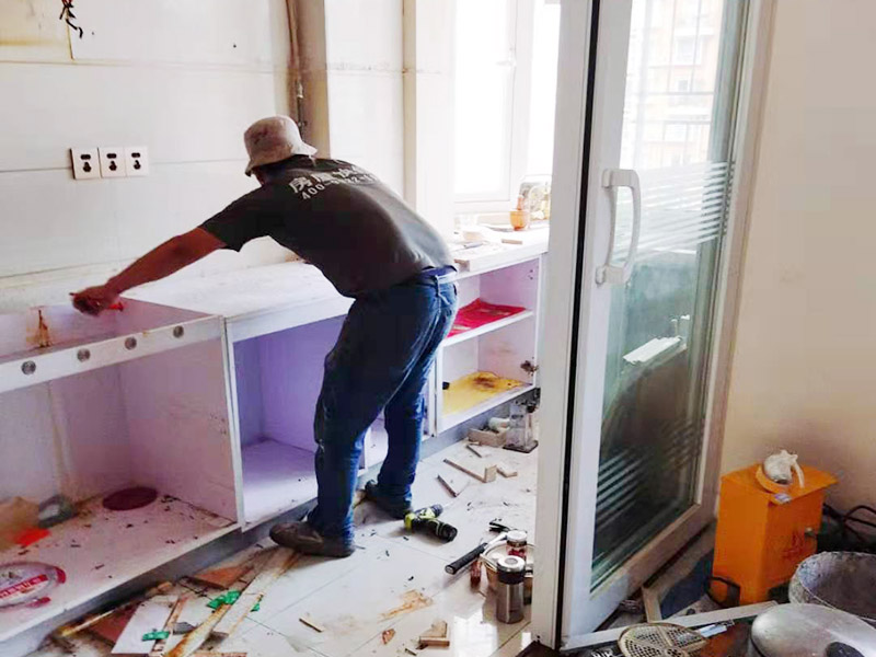 廚房櫥柜安裝：清理：對廚房作防護措施，掛擋布、廚房墻面等做簡單清理，以便準確測量地面水平。櫥柜安裝一般先從地柜開始。把櫥柜外包裝拆開后。在櫥柜的邊緣貼上防火膜封邊。在下柜底部安裝上支腳，根據地面水平高度調整櫥柜。 櫥柜安裝需要找出基準點，L型廚房從直角處向兩邊延伸，U型廚房則先將中間的一字型柜體放整齊后，再從兩個直角處向兩邊站看，這樣可以避免出現縫隙。櫥柜框架安裝牢固定位后，需要在櫥柜背面安裝背板。一般安裝下水都會現場開孔，工人會用專業的打孔器按照管道大小進行打孔，打孔的直徑應比管道大3-4毫米，并且打孔后要將開孔部分用密封條封邊，防止邊緣滲水木材變形。